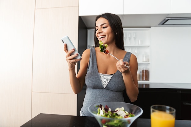 Healthy pregnant woman indoors at home at the kitchen using mobile phone eat salad.