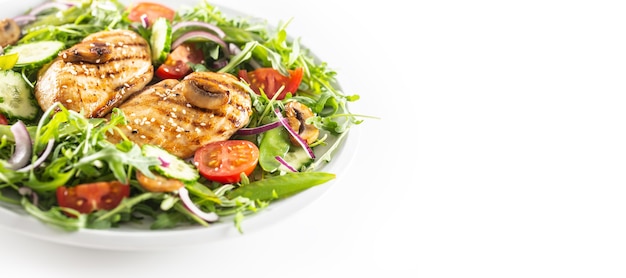 Healthy salad with grilled chicken topped with sesame seeds, rocket, spring peas, cherry tomatoes, sliced cucumber, grilled mushrooms and red onion, isolated on left half of the white background.