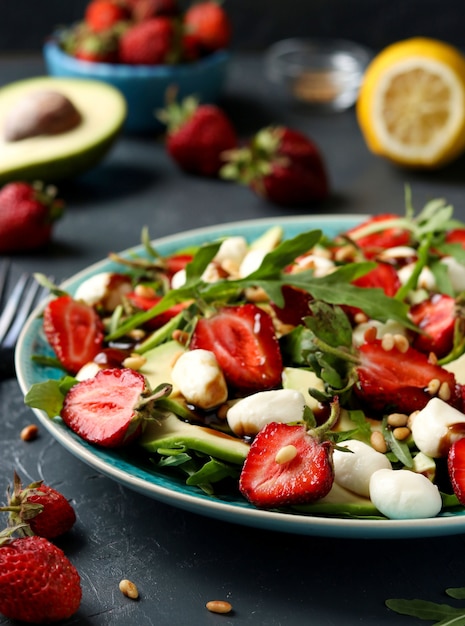 Healthy salad with strawberries, avocado, arugula and mozzarella, dressed with olive oil and balsamic dressing located in a plate on a dark 