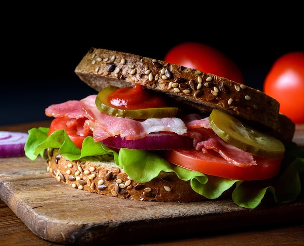 Healthy sandwich freshly prepared with bacon, tomato, cucumber, onion on whole grain wheat toast on dark background.