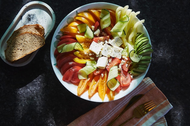 Healthy seasonal salad with red orange and yellow tomatoes sweet pepper avocado cucumbers feta cheese sesame on dark marble background close up Natural organic food healthy dinner