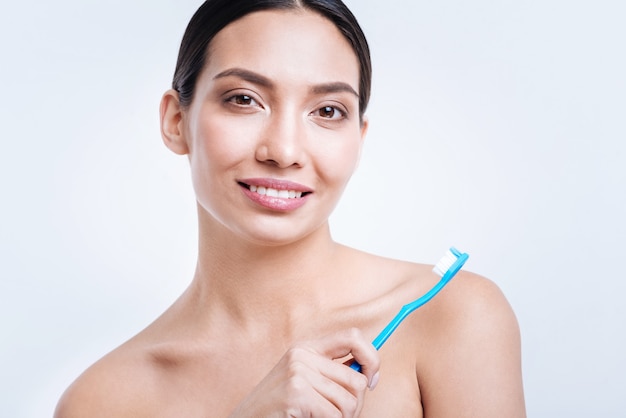 Healthy smile. Pleasant upbeat young woman showing her green toothbrush and smiling brightly while posing against a white wall