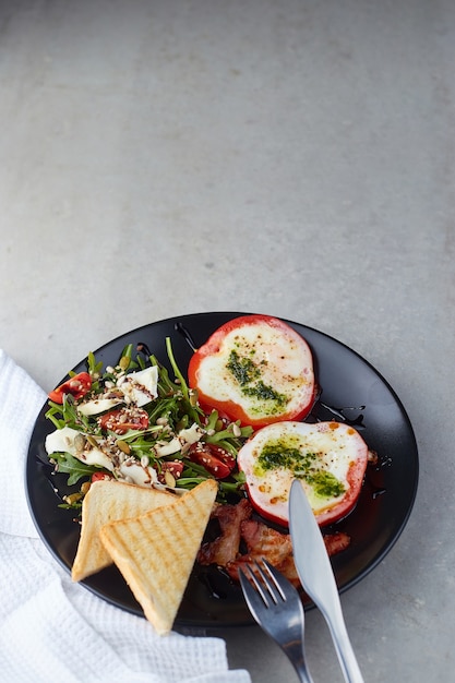 Healthy and tasty breakfast. Fried Eggs in Red Pepper rings, bacon and salad with arugula, cherry tomatoes, seeds and hard cheese on black plate