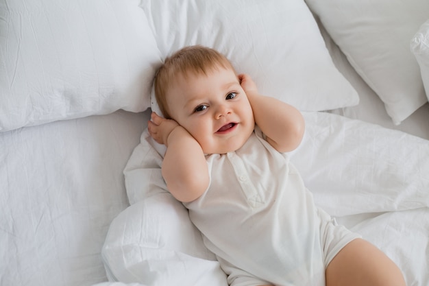 Healthy toddler boy smiles in white bodysuit lies in bed with white linen