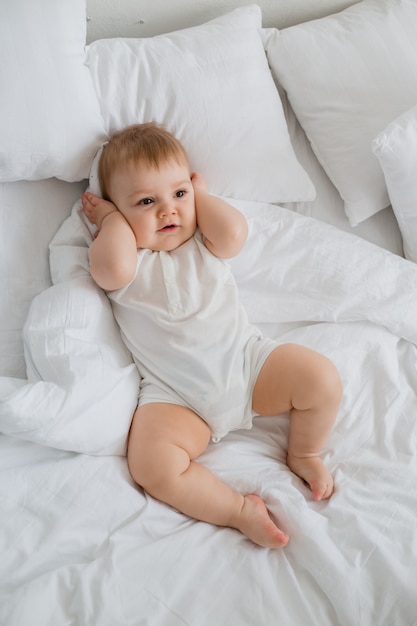 Healthy toddler boy smiles in white bodysuit lies in bed with white linen