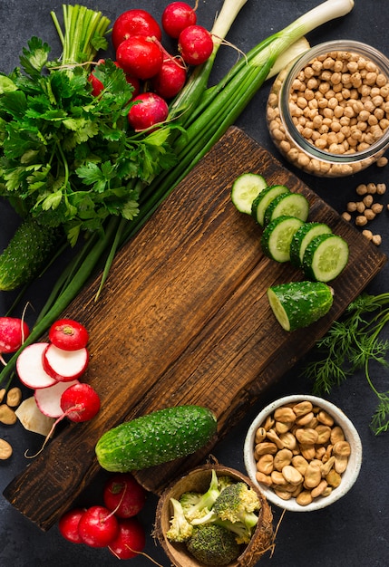 Healthy vegan food veggie cooking concept. Wooden cutting kitchen board with fresh vegetables, herbs and cereal on dark background top view