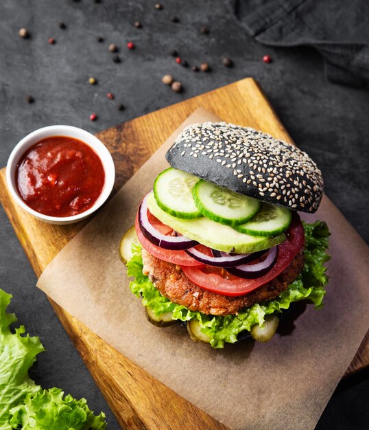 Healthy vegetarian burger with black bun chickpeas cutlet salad cucumber on a cutting board with tomato sauce and spices on dark background The concept of vegan fast food