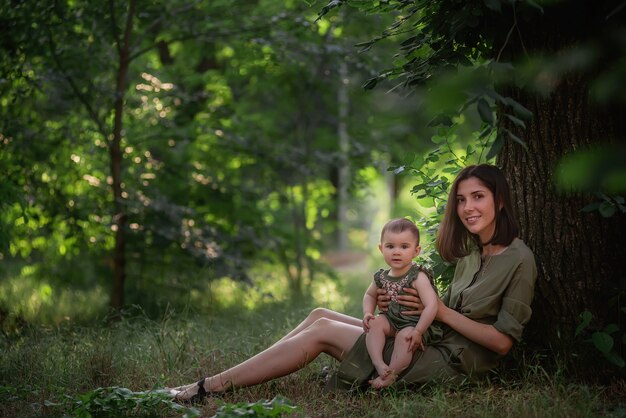 A healthy young mother holds a toddler baby in her arms. A happy family sits on the green grass, under a tall tree, plays, hugs