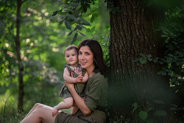 A healthy young mother holds a toddler baby in her arms. A happy family sits on the green grass, under a tall tree, plays, hugs