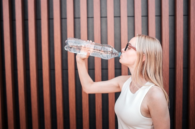 Healthy young woman is drinking water from bottle. She is standing at the wall. Isolated on striped background