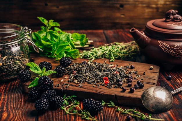 Heap of Dry Green Tea and Fresh Blackberries on Wooden Cutting Board. Bundles of Mint and Thyme Leaves. Clay Kettle.