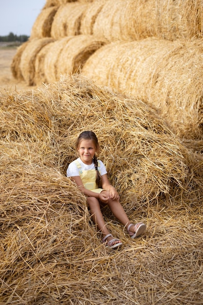 Heap of hay on field with little girl sitting in it smiling with sly pensive eyes looking away holding hay in hand wearing sundress Having fun away from city on field full of golden hay