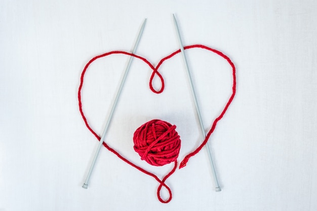 Heart made of thread with woolen yarn and knitting needles on white wooden background. 