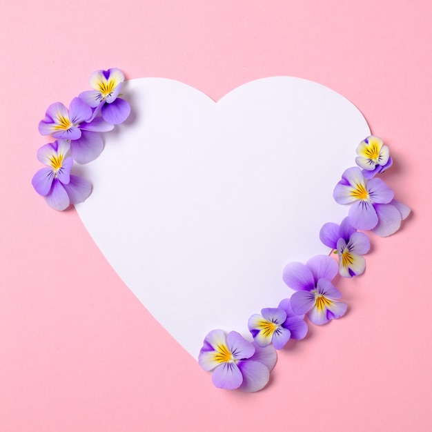 Heart-shaped blank paper card and wild violet flower petals on pastel pink background. frame