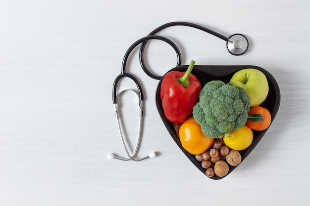 Heart shaped bowl with vegetables fruits nuts and stethoscope Healthy eating concept