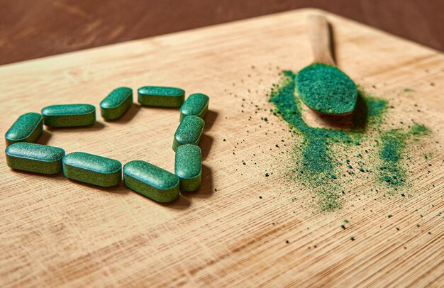 Heart shaped chlorella or spirulina tablets and powder in wooden spoon on light wooden background