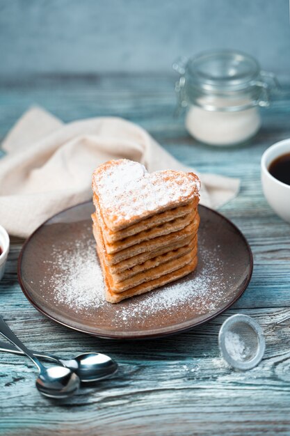 Heart of waffles sprinkled with powdered sugar in a brown plate on a wooden gray-blue background.