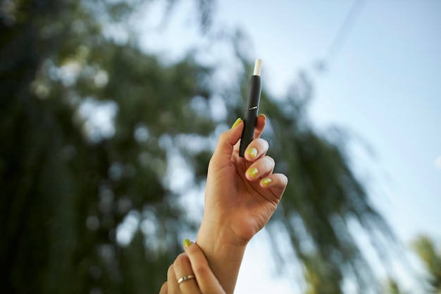 Heating tobacco system IQOS on summer background. Woman hand holding a e-cigarette.