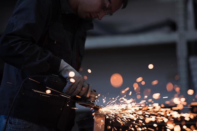 Photo heavy industry engineering factory interior with industrial worker using angle grinder and cutting a metal tube contractor in safety uniform and hard hat manufacturing metal structures