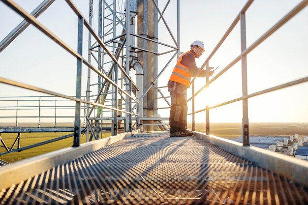 A heavy industry worker is using tablet while leaning on railing on high metal construction