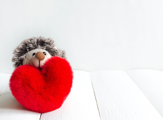 Hedgehog toy and red soft heart on a white wooden background for valentine day with copy space