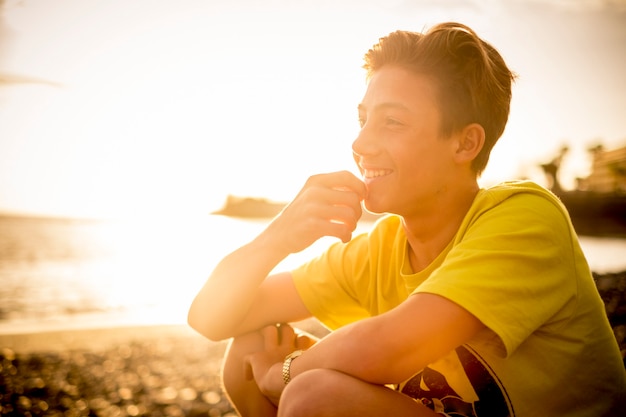 heerful handsome young boy sitting at the beach smiling and looking at the horizon - happy people in outdoor leisure vacation activity during golden sunset