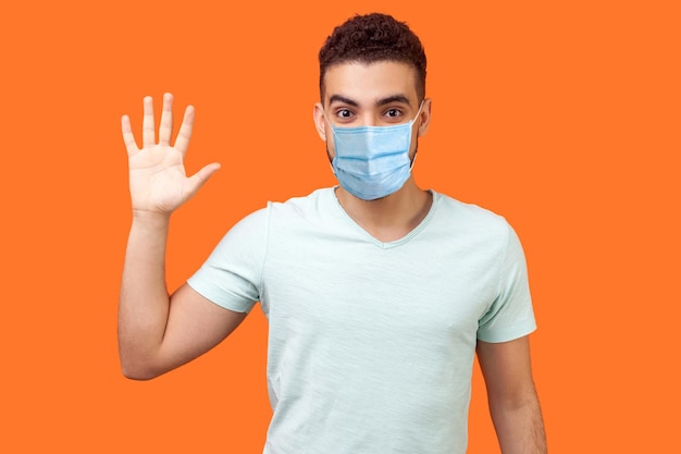 Hello Portrait of friendly glad handsome man with medical mask in casual white tshirt showing hi gesture with waving hand and smiling sincerely indoor studio shot isolated on orange background