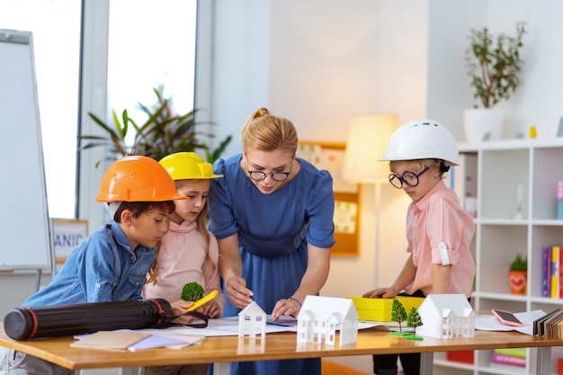 Helpful teacher. Stylish teacher helping her pupils in helmets making construction sketches at lesson