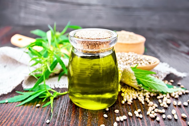 Hemp oil in a glass jar, grain in a spoon and flour in a bowl on a napkin of burlap, leaves and stalks of cannabis on a wooden board background