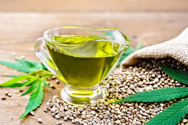 Hemp oil in a glass sauce-pot with grain in a bag, leaves and stalks of cannabis on a wooden board background