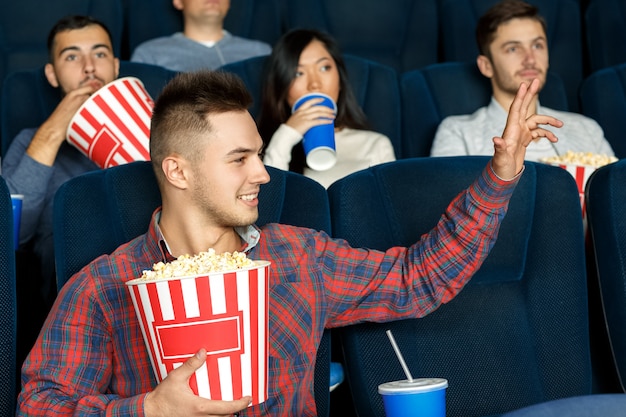 Over here. Young casual man waving his hand to the friend while sitting in the cinema enjoying movies