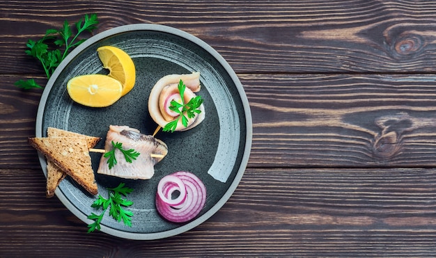 Photo herring roll on a skewer with parsley, onion and lemon wedge, top view. the sandwiches are arranged on a plate, on a dark wooden table with space for text.fast food or snack