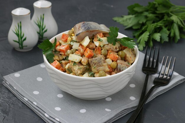 Herring salad with carrots, eggs and peas in a white bowl on a dark background