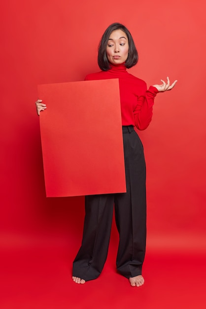 Hesitant Asian woman looks with doubt at billboard shrugs shoulders doesnt know what information to place wears turtleneck black trousers isolated over bright red wall