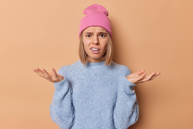 Photo hesitant clueless blonde woman spreads palms feels confused and bewildered frowns face wears pink hat casual blue jumper cannot make decision isolated over brown background. human reactions.