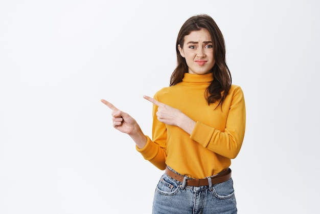 Hesitant and unsure young girl grimacing and frowning pointing fingers left with doubtful expression feeling uncertain telling bad news standing on white background