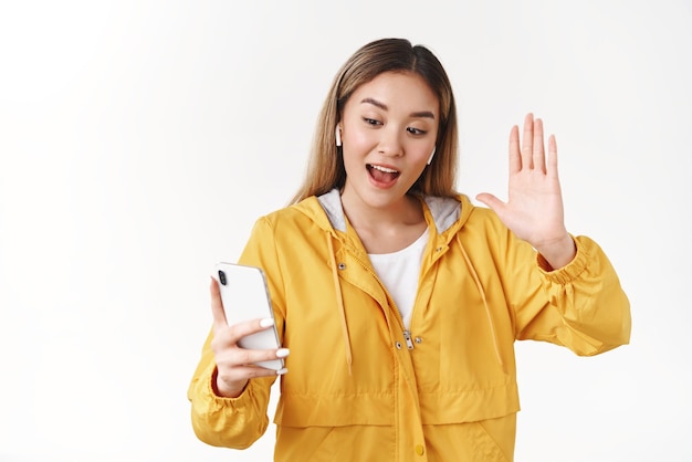 Hey good finally see you Joyful stylish cool modern asian hipster girl blond hairstyle raise hand whats up hello gesture wear wireless earbuds hold smartphone look screen talking videocall