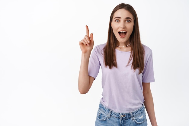 Hey look Excited girl turns your attention to awesome promo deal showing best discounts in store pointing finger up and stare impressed at camera white background