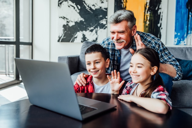 Hey there. Positive minded retired man and his little grandchildren smiling and waving their hands while having a video call at home.