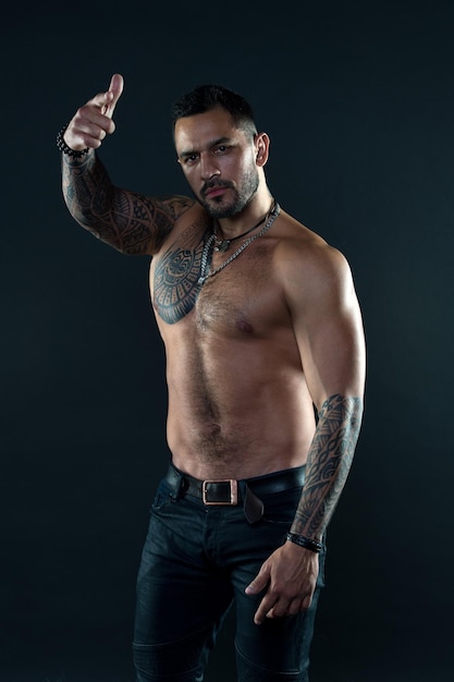 Hey you Handsome fit tattooed man wear jeans pointing forward Man handsome shirtless muscular with jeans over dark background Muscular tattooed athlete look attractive Sport and fashion concept