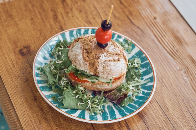 High angle of appetizing vegetable burger with tomatoes olives cucumbers and lettuce served on colorful plate on wooden table