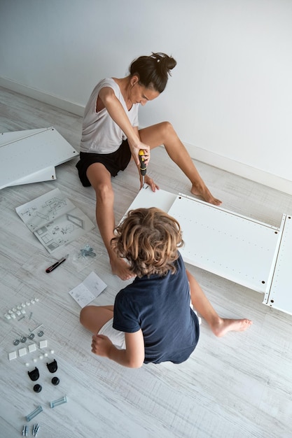 High angle of concentrated woman tightening screws with screwdriver while preteen boy reading instruction and helping in living room at home