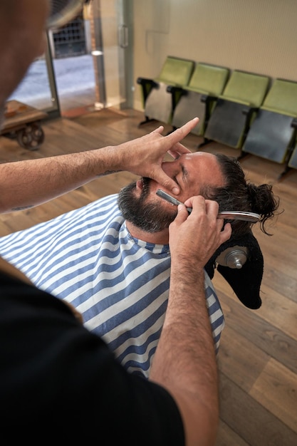 High angle of crop anonymous barber using straight blade to cut beard of male client while working in barbershop