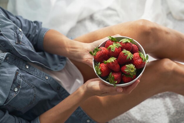 Photo high angle of crop female holding ceramic bowl with appetizing strawberries while sitting on white fabric in light room