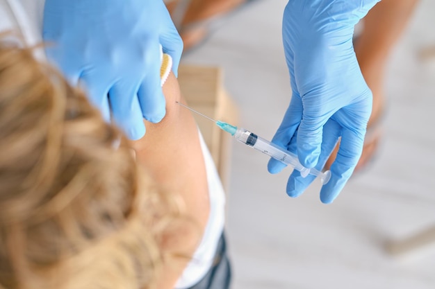 High angle of crop unrecognizable female medic in latex gloves injecting coronavirus vaccine into shoulder of patient in clinic