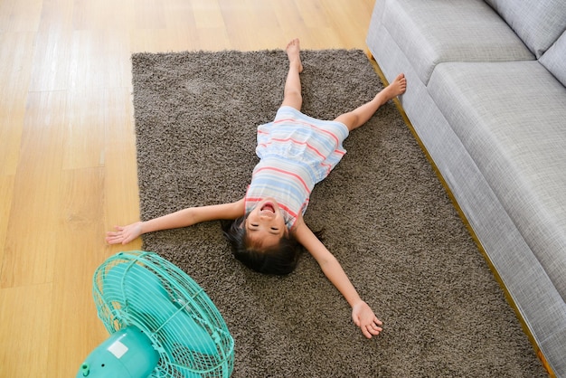 high angle photo of happy cute little girl lying down on floor and enjoying electric fan blowing cool wind in summer day.