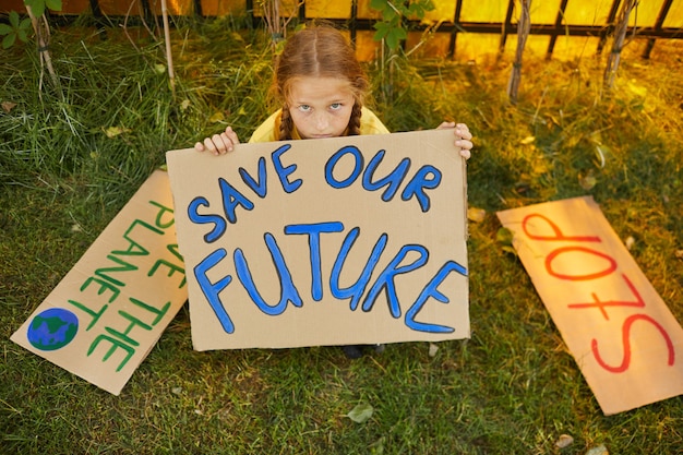 High angle portrait of freckled girl holding SAVE OUR FUTURE sign and looking up at camera while protesting for nature sitting on grass outdoors, copy space