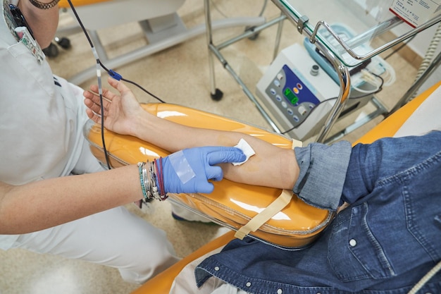 High angle of professional female medical worker in white uniform and blue gloves putting white sterile cotton on arm of patient in jeans jacket