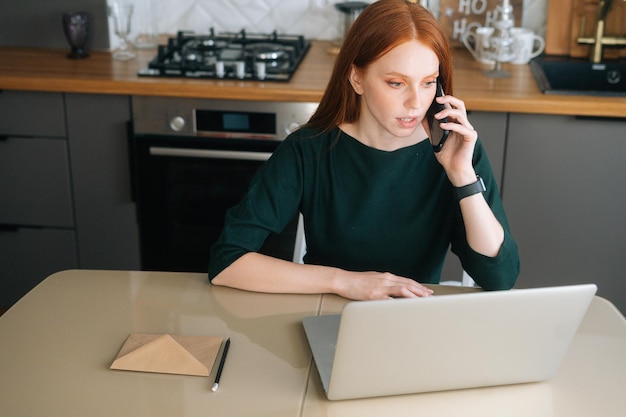 High-angle view of angry female freelancer talking on mobile phone and using laptop sitting at table in kitchen with modern interior. Annoyed young woman talking on cellphone with support manager.