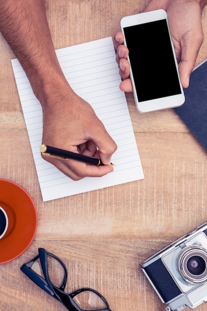 High angle view of businessman hand holding smart phone while writing on notepad by camera and coffee on table